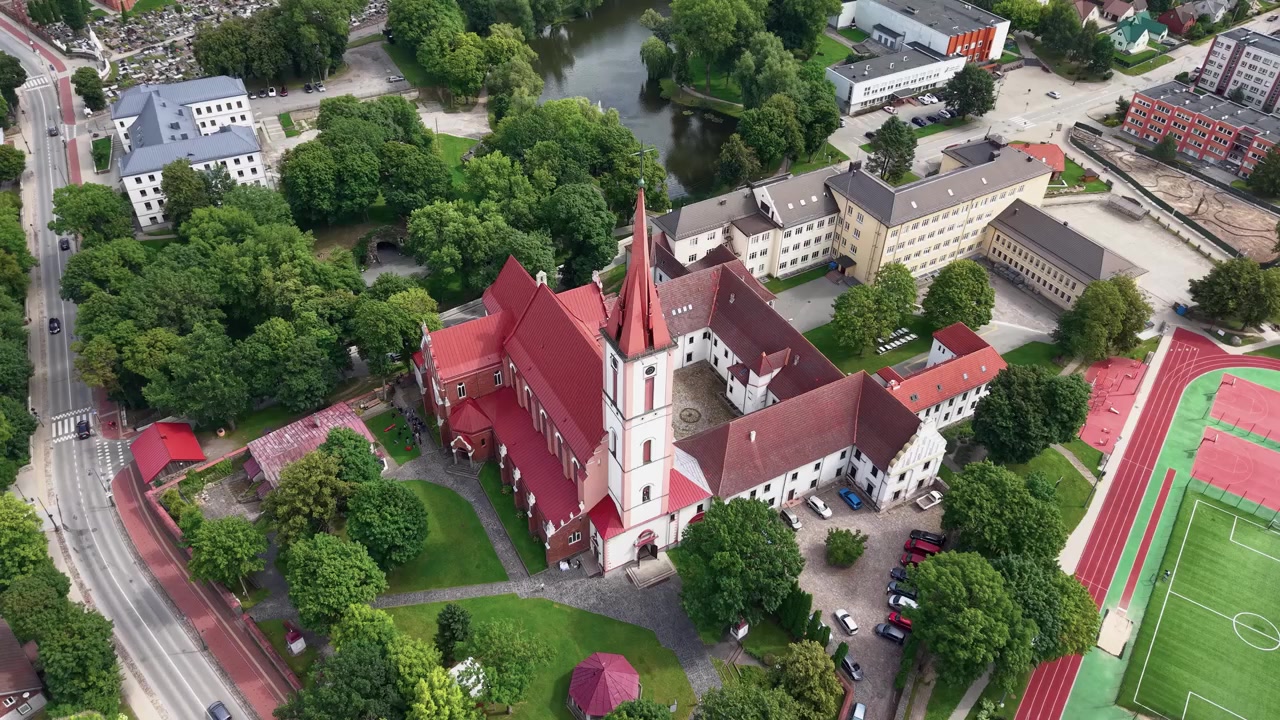 Imponerende dronevideo viser Kirken for Herrens Åbenbaring til Jomfru Maria i Kretinga, Litauen, omgivet af frodigt grønt, historiske bygninger og et sportsstadion i dagslys