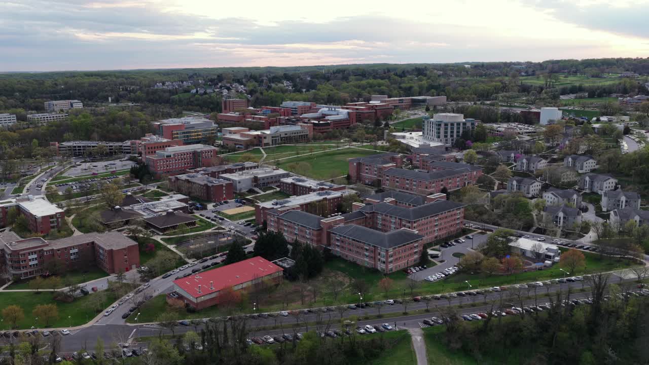 Quartiere suburbano di Baltimora che conduce verso lo skyline del centro, con la strada principale che attraversa le aree residenziali sotto soffici nuvole primaverili e un'atmosfera urbana calma. Ampia ripresa aerea laterale.
