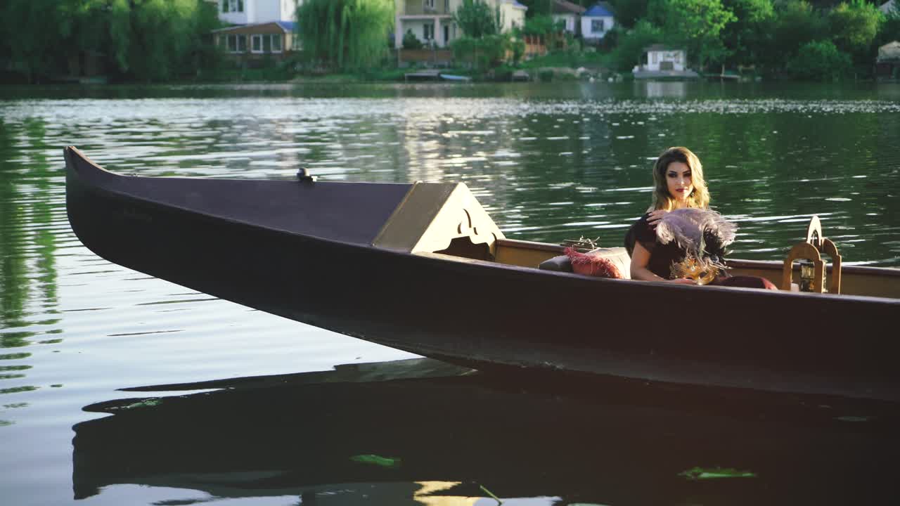 Young beautiful woman in boat is holding the feather and showing her shoulder on the river background. Sexy female with long dark hair floating in summer