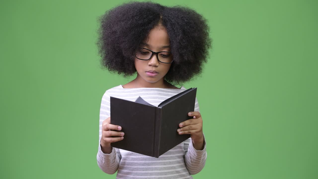Young cute African girl with Afro hair studying against green background
