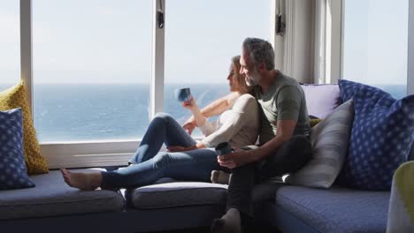 Happy-caucasian-mature-couple-drinking-coffee-and-talking-in-sunny-living-room