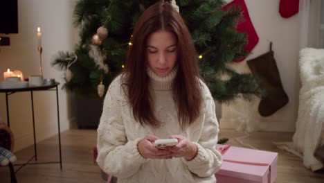 A-brunette-girl-in-white-light-looks-and-does-something-on-her-white-phone-while-sitting-near-a-large-pink-gift-and-a-New-Year-tree-at-home-in-a-cozy-apartment-decorated-in-the-style-of-the-New-Year