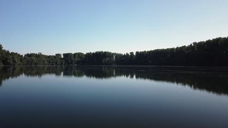 Drone-flying-close-over-the-water-of-a-dark-eerie-lake-with-reflections-of-trees-and-blue-skies-in-Europe