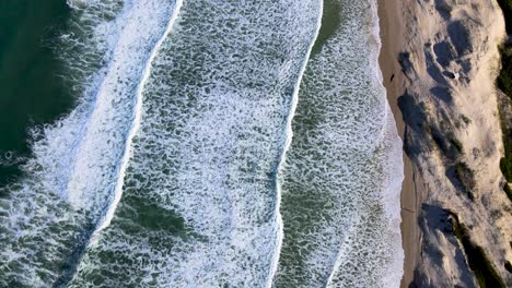 Drone-view-of-beautiful-turquiose-sea-waves-breaking-on-sandy-coastline-meeting-deep-blue-ocean-water-and-foamy-waves