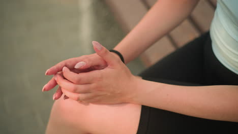 incomplete view of lady seated on wooden bench outdoors with leg crossed, gently rubbing her hands together resting on knee, background softly blurred with warm sunlight