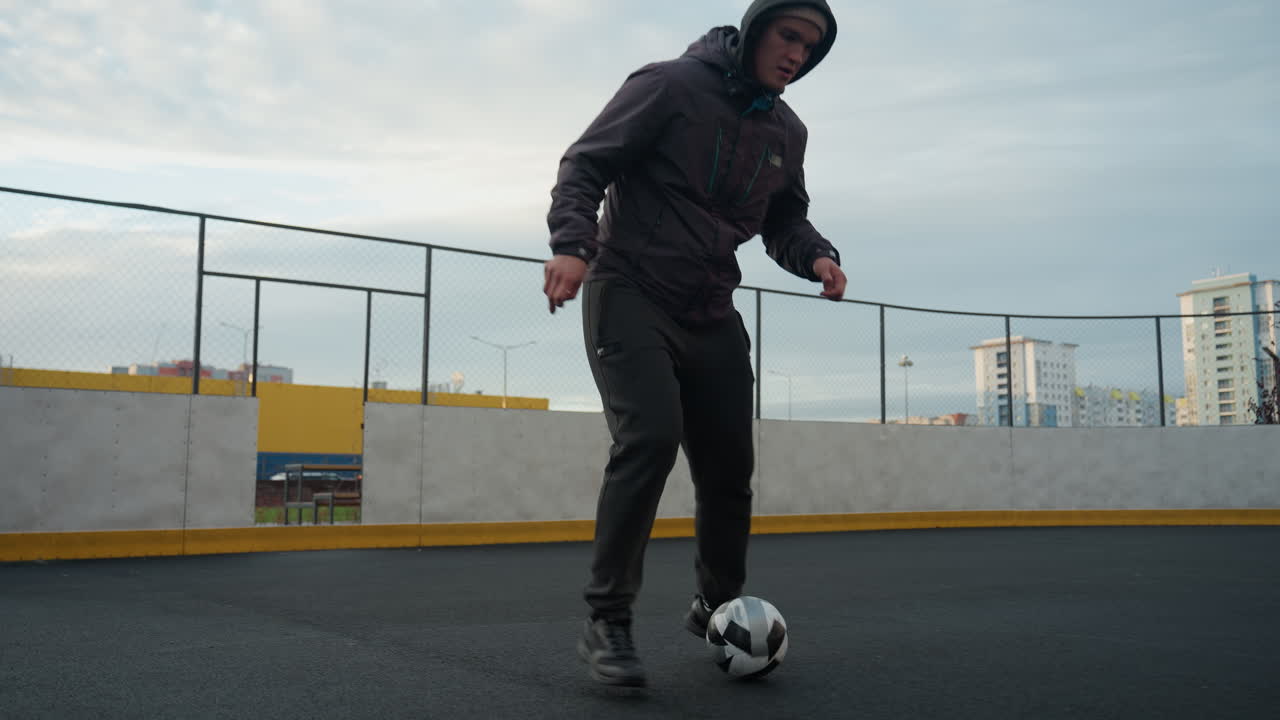 Young athlete skillfully training with soccer ball on sport arena during a practice session, background includes urban buildings, fencing, and goalpost