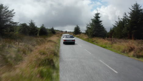 Forwards-tracking-of-vintage-sports-car-on-road-in-highlands.-Meadows-and-trees-in-forest-around.-Overcast-sky.-Ireland