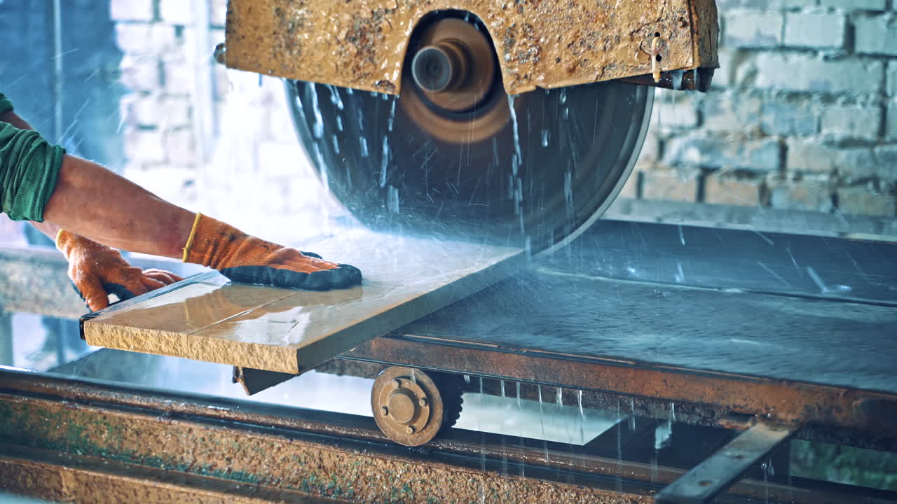 Male's hands in gloves holding stone slab close to the automated machine. Automated circular saw cuts the stone slab at industrial factory. Worker controls the electric cutter machine.