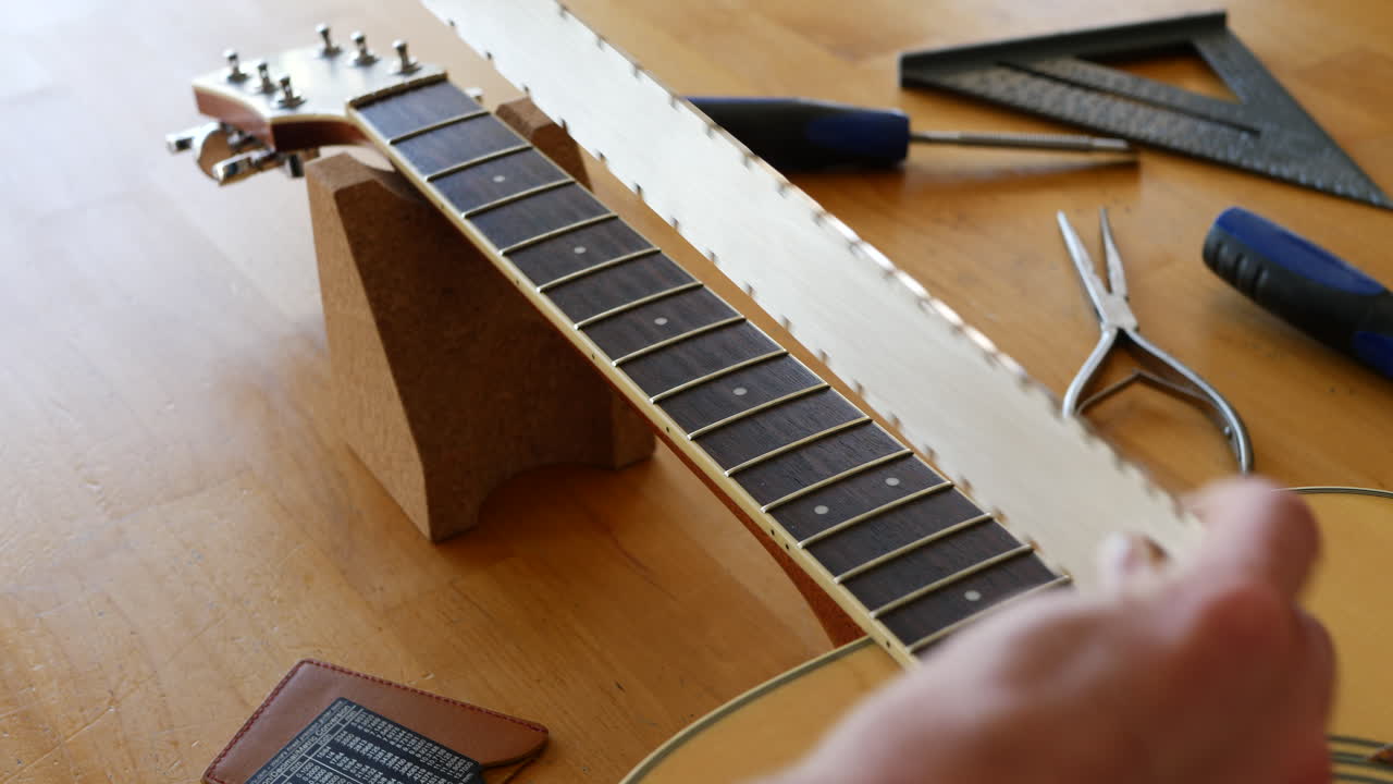 Hands of a luthier craftsman measuring and leveling an acoustic guitar neck and fretboard on a wood workshop bench with lutherie tools