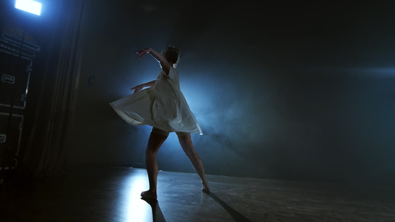 The zoom camera moves the woman dancer across the stage with software and smoke. A modern dramatic ballet a woman in a white dress spins on one leg and jumps..