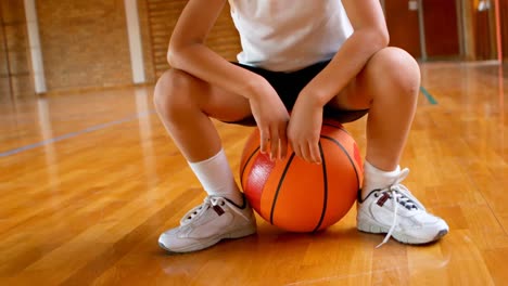 athletic african american schoolboy sitting on basketball in basketball court at school 4k