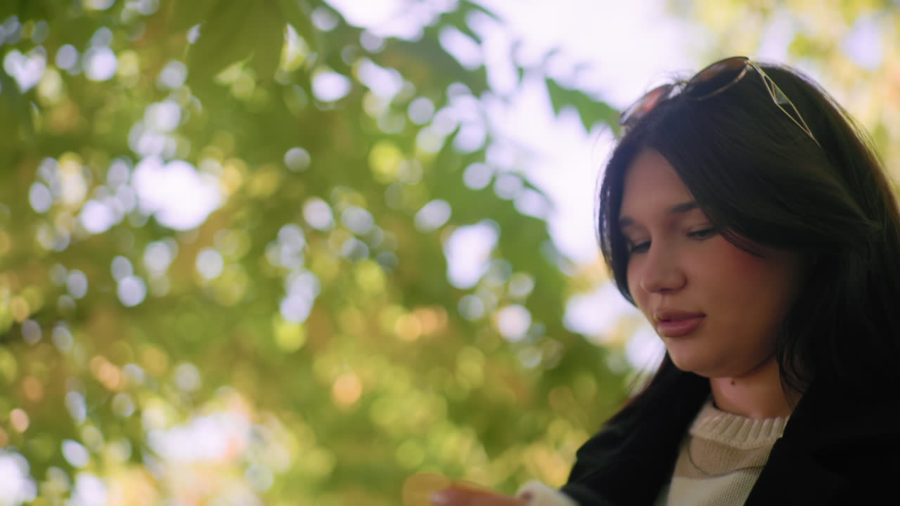 Side view of smiling lady in black coat raises hand toward dry leaf on tree, admires detail, soft autumn bokeh around, gentle sunlight through branches, serene moment of curiosity in quiet park