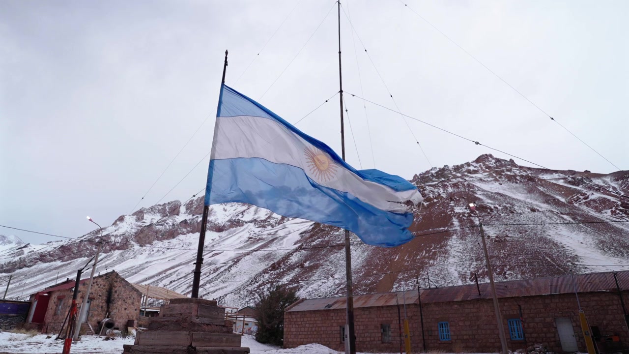 Grand drapeau argentin flottant sur un grand mât devant les sommets enneigés des Andes à Puente del Inca. Vue statique grand-angle au ralenti par temps nuageux