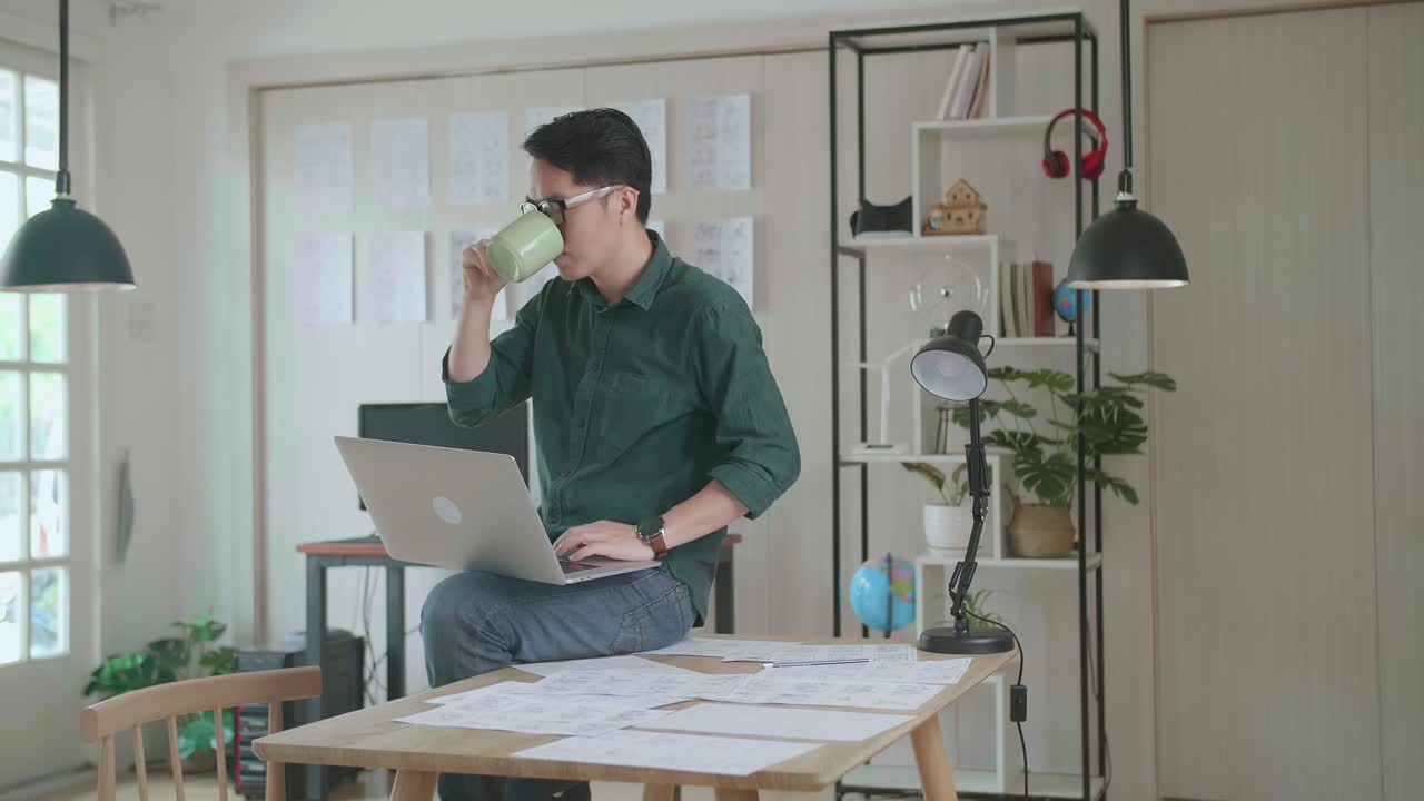 Creative Designer Sits On His Desk Holds Laptop On The Knees Drinks Coffee While Working On The Project. Creative Designe And Gaming Content Development Studio