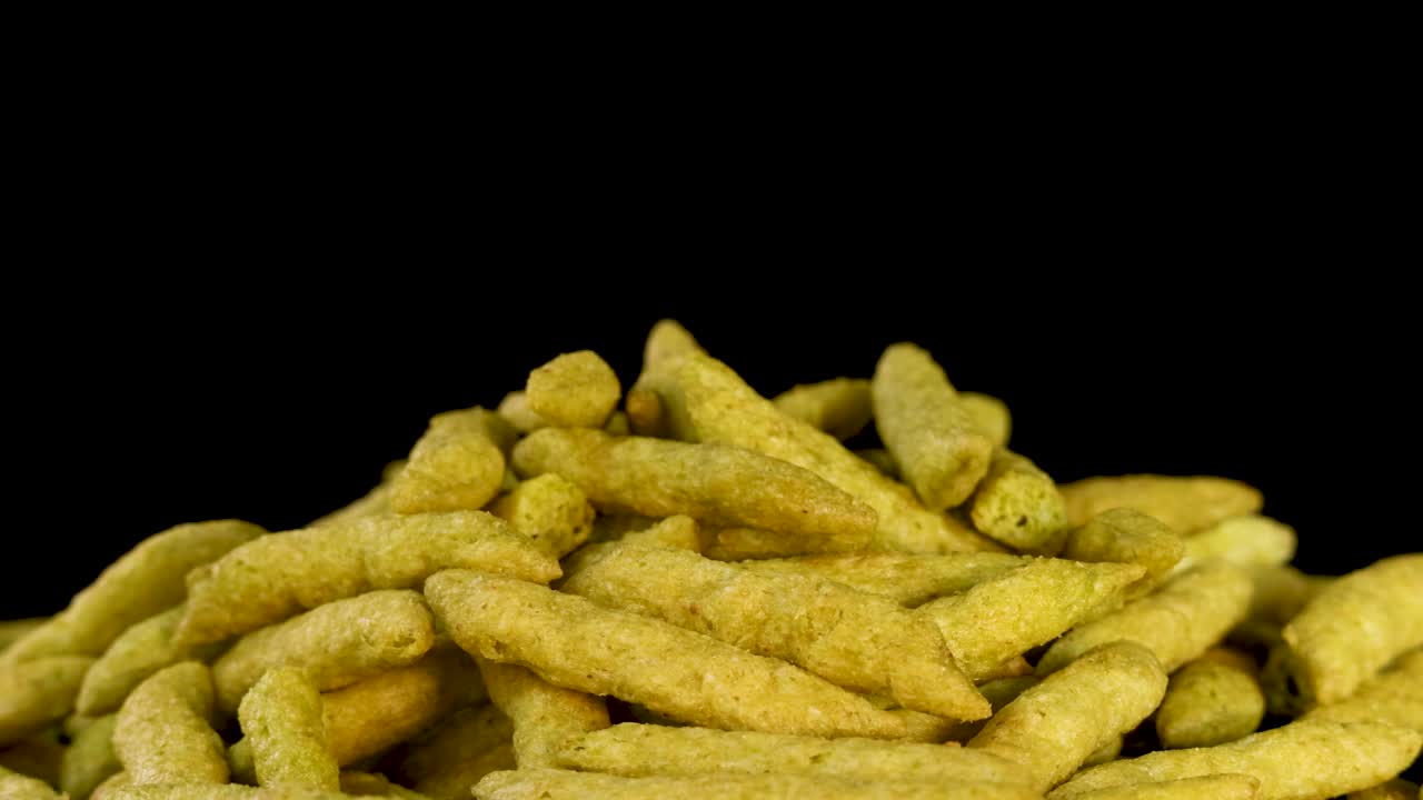 Golden puffed green pea snack sticks rotate under studio lighting against a clean black background