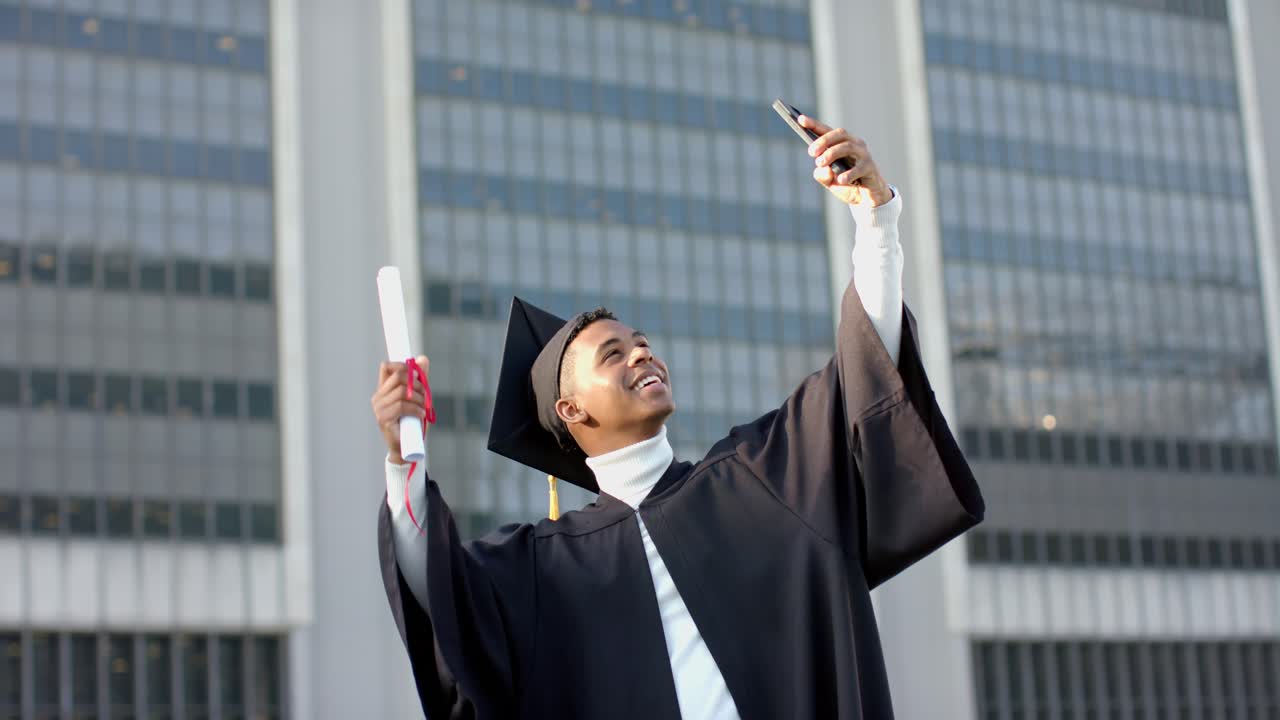 After graduating, man in gown holding diploma lifting smartphone for graduation selfie at plaza