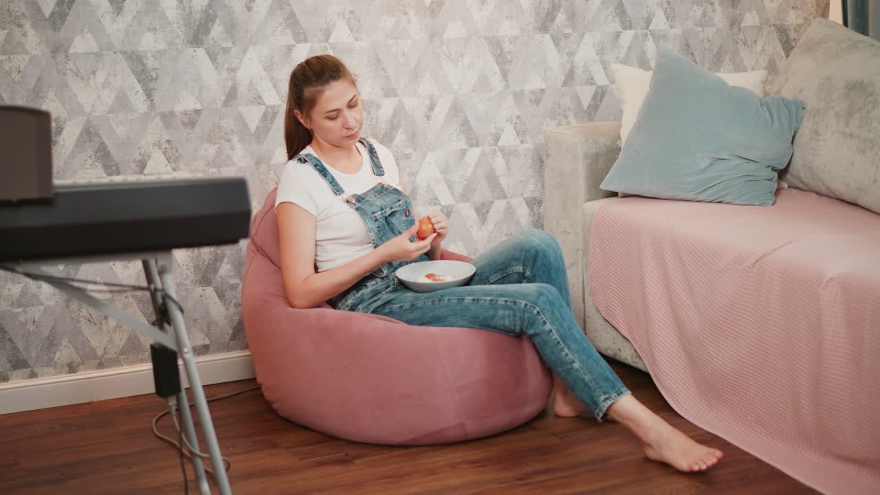 Relaxed woman in denim overalls seated on soft beanbag carefully removing egg shell into bowl on lap, with digital piano visible in background and cozy interior featuring pastel furnishings