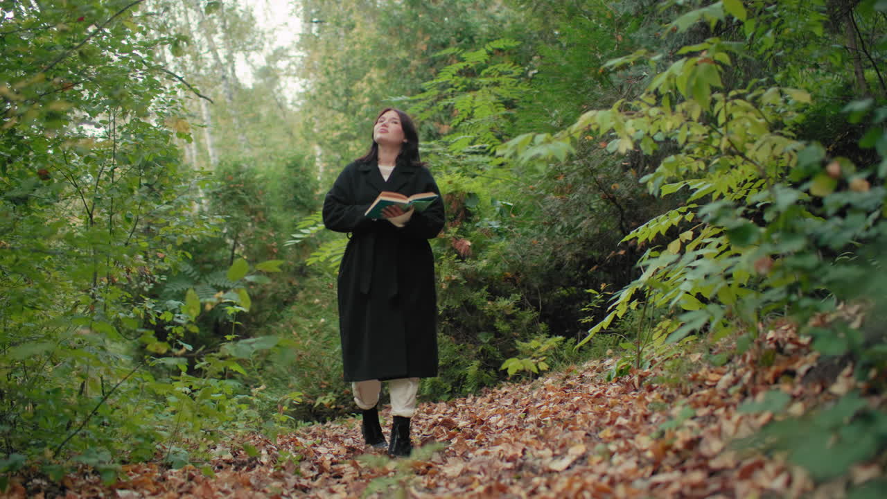 Persona silenciosa camina con un libro en las manos y hojea sus páginas mientras pasea por el bosque, disfrutando de la naturaleza y del fresco aire otoñal, con un humor tranquilo y pasos lentos y conscientes por el sendero lleno de hojas, sintiendo curiosidad, leyendo para evadirse en medio del follaje verde