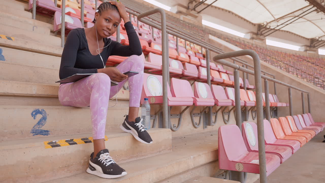 Mujer joven de raza negra sentada en las escaleras del estadio revisando su teléfono, con postura relajada, asientos rosas vacíos, atuendo casual con pantalones rosas, zapatillas negras, barandilla de metal, recinto deportivo urbano, estado de ánimo introspectivo.