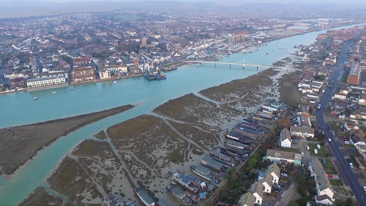 Filmagem de drone com panorâmica ascendente mostrando um rio e casas flutuantes cercados por uma cidade costeira em Brighton, Inglaterra, ao pôr do sol.
