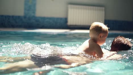young man rowing under the water with his blonde son on the back. little boy is above the water. indoors swimming pool. slow motion
