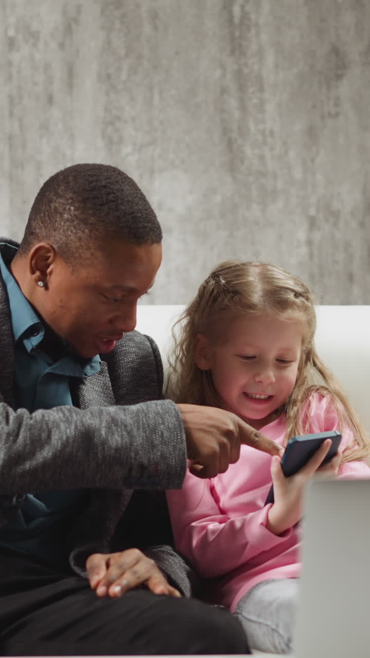 Careful African-American private English teacher talks to funny little schoolgirl holding phone at break during tuition in living room slow motion