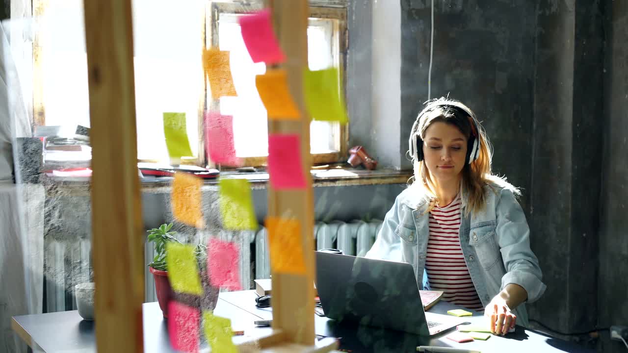 En kreativ ung forretningskvinde lytter til musik i høretelefoner og danser, mens hun arbejder ved skrivebordet med sin bærbare computer på et moderne kontor. Et glas med farvede klistermærker er i forgrunden.