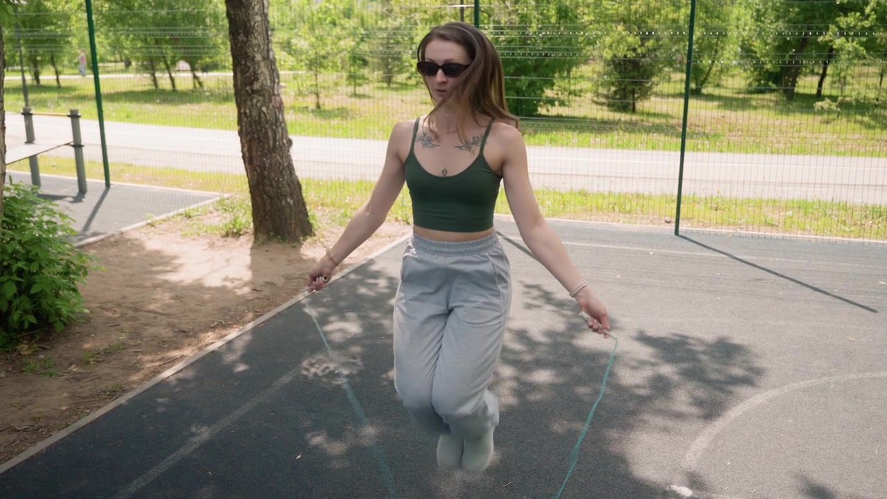 Close up of young woman actively skipping rope in outdoor park setting wearing sunglasses and casual athletic clothing. background features greenery trees exercise equipment other people
