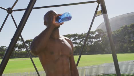 Fit-shirtless-african-american-man-exercising-on-climbing-frame-outdoors,-resting-and-drinking-water