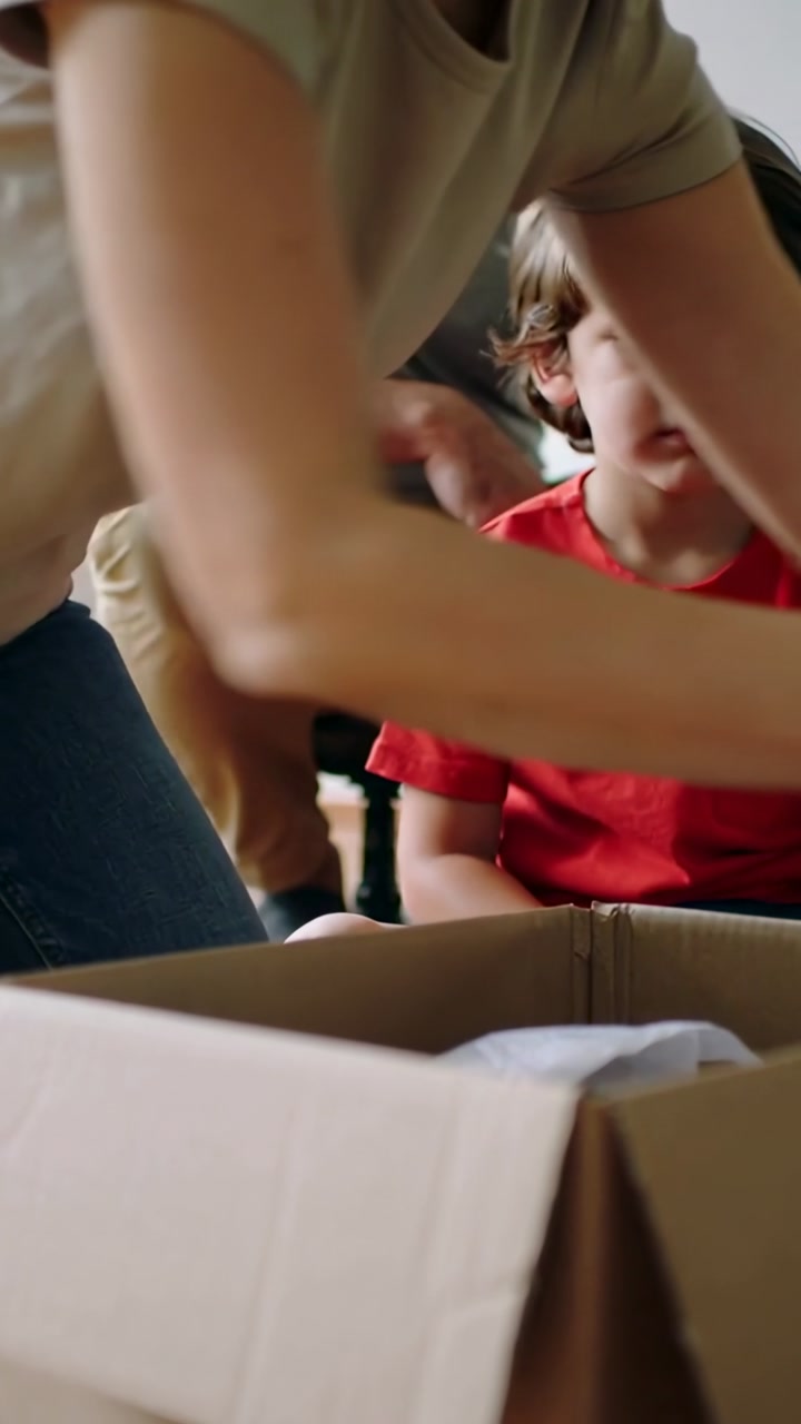 Vertical video: Unpacking adult reaching into box at home after delivery, child in red tee watching