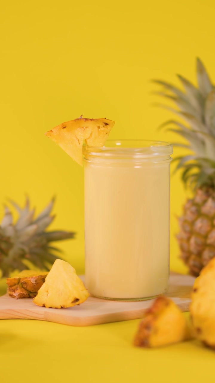 Una mano posiziona una fetta d'ananas su un bicchiere di frullato cremoso, circondato da frutta fresca, su uno sfondo giallo brillante.