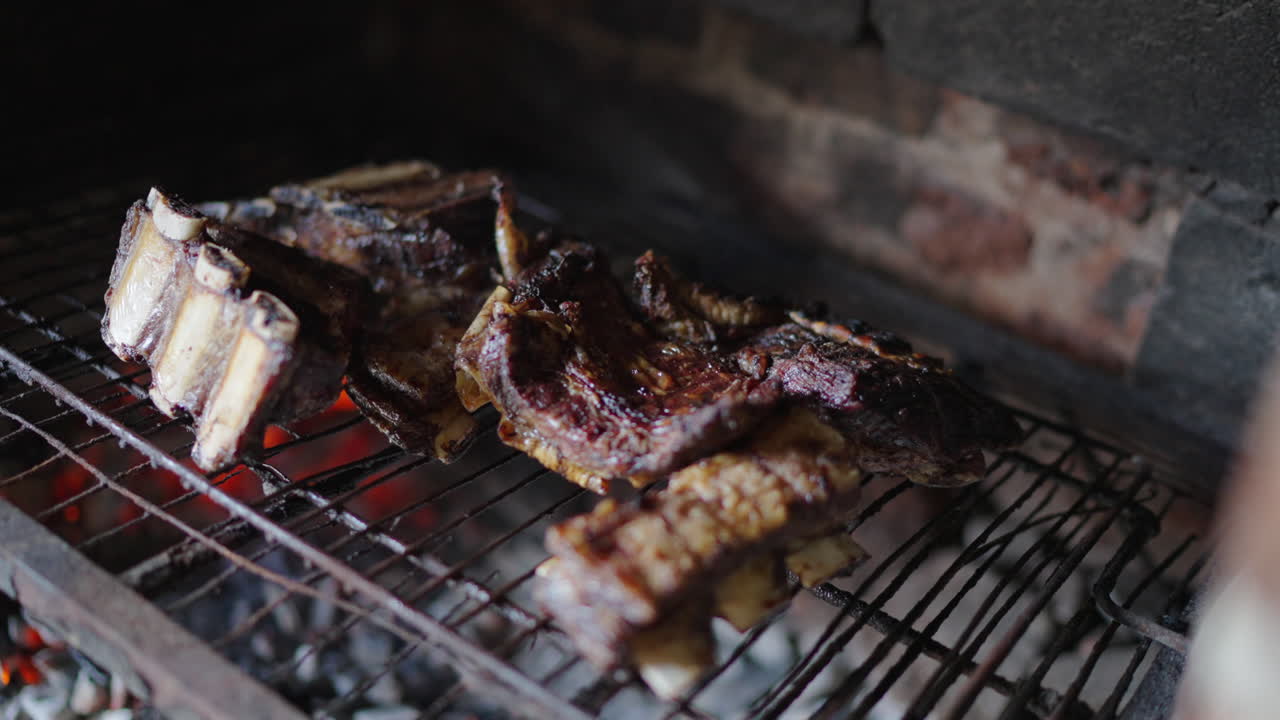 Close up of traditional Argentine asado ribs cooking over glowing wood embers on a metal grill inside a rustic brick parrilla, with visible smoke, caramelized crust, and rendered fat, slow motion.