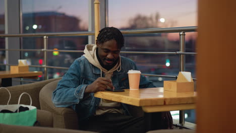 young man in denim jacket and hoodie seated in cozy cafe during sunset, gently smiling while holding coffee cup, surrounded by warm indoor lighting, soft armchair, napkin box, and paper shopping bag