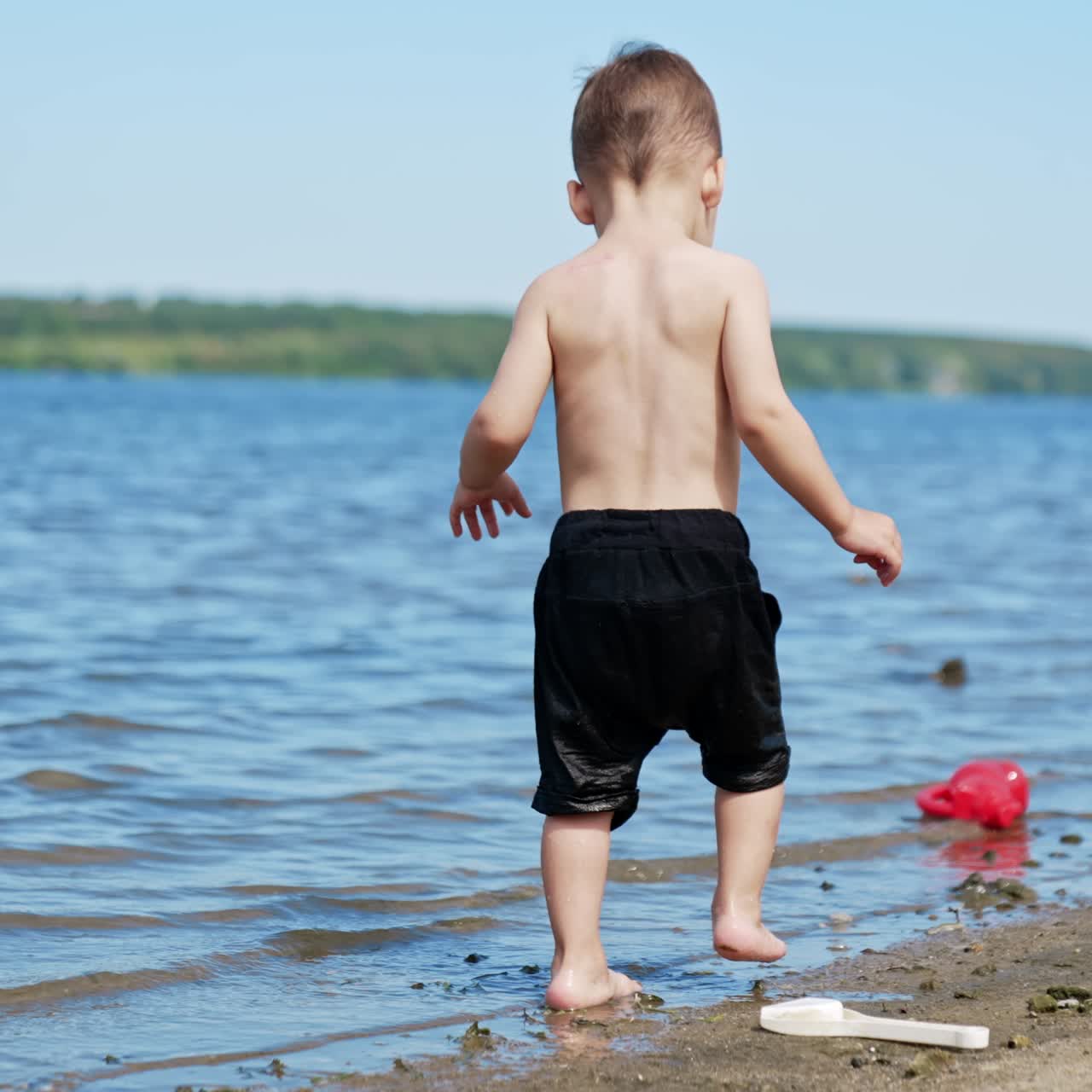 Cute kid in black shorts carefully steps by the water and sand in the beach. Caucasian child having rest near the river in summer