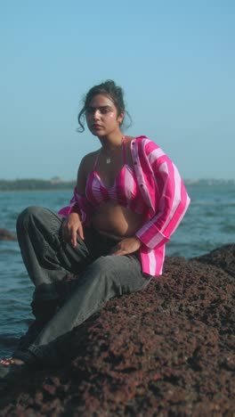 Young woman sitting on a rock at the beach, wearing pink striped top and jeans