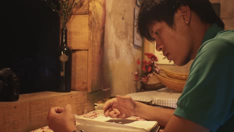 young asian male reading a book at a desk in a rustic room