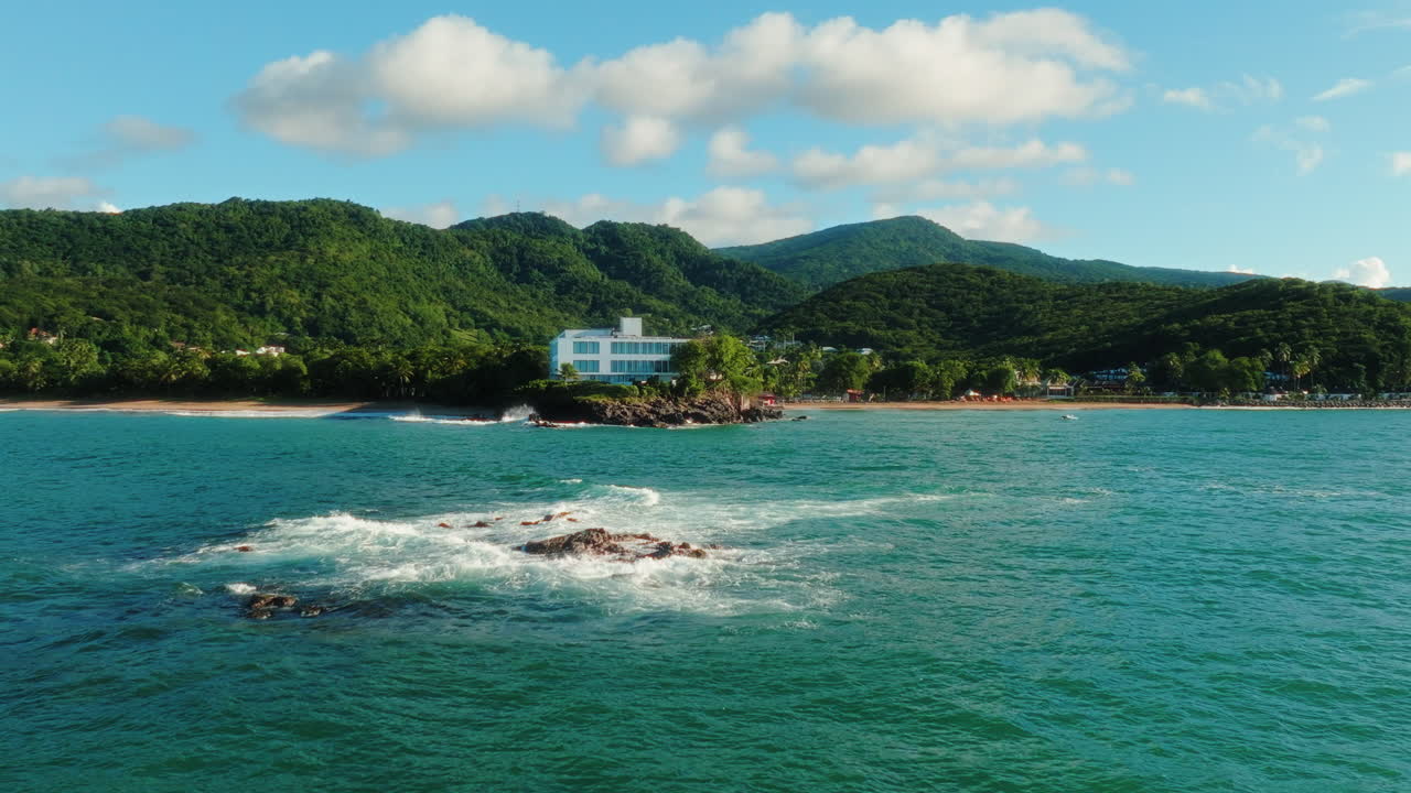 Drone flies backward above the turquoise sea in Guadeloupe, revealing waves crashing on rocks and a large hotel complex on the lush tropical coast