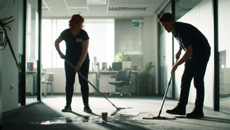 two office cleaners collaboratively addressing a spill on the carpet with mops during a transitional work environment in an office setting