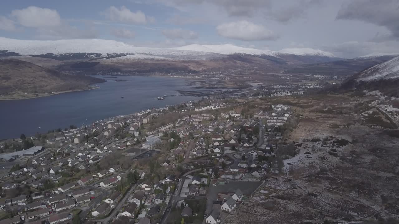 Aerial drone footage of Fort William in the Scottish Highlands on a bright, sunny winter day, showing a scenic mountain landscape with snow-covered peaks and a clear blue sky.
