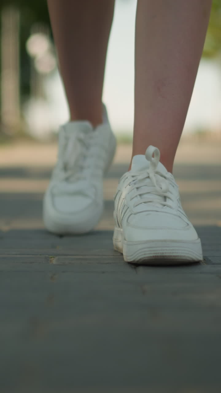 Close-up leg view of individual in white sneakers walking on interlocked pathway under warm sunlight, with blurred background featuring tall trees, lush greenery, and serene outdoor setting