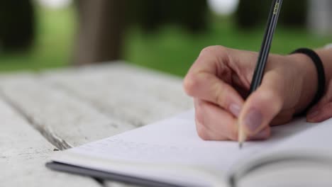 Primer-Plano-De-Manos-Femeninas-Escribiendo-Recuerdos-En-El-Diario,-Sentado-En-Una-Mesa-Blanca-En-El-Jardín-Verde,-Rack-Fokus