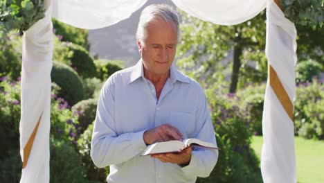 smiling caucasian senior male wedding officiant standing in outdoor altar, holding book