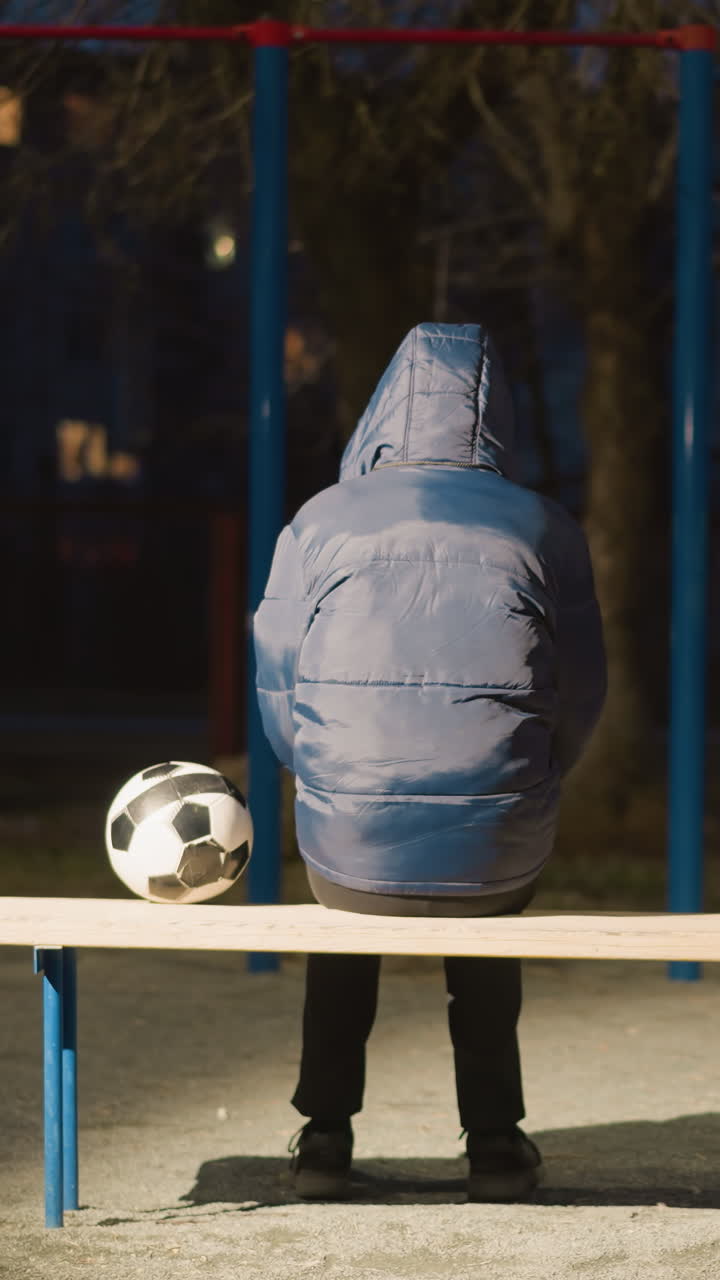 задний вид человека в синем куртке с капюшоном, сидящего на скамейке на открытом воздухе ночью с футбольным мячом, помещенным рядом с ним, с легким отражением от окружающих