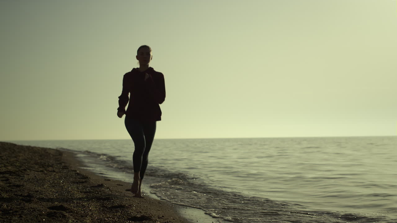 Siluet wanita berlari di pantai saat matahari terbenam. Gadis jogging di tepi laut pada sore musim panas.
