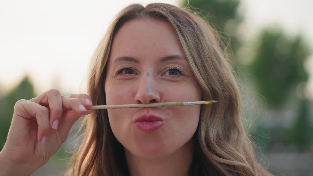playful woman in summer park holds paintbrush under nose sniffing it gently paint dots on face smiling with trees and pool reflection blurred behind under evening light capturing candid art moment