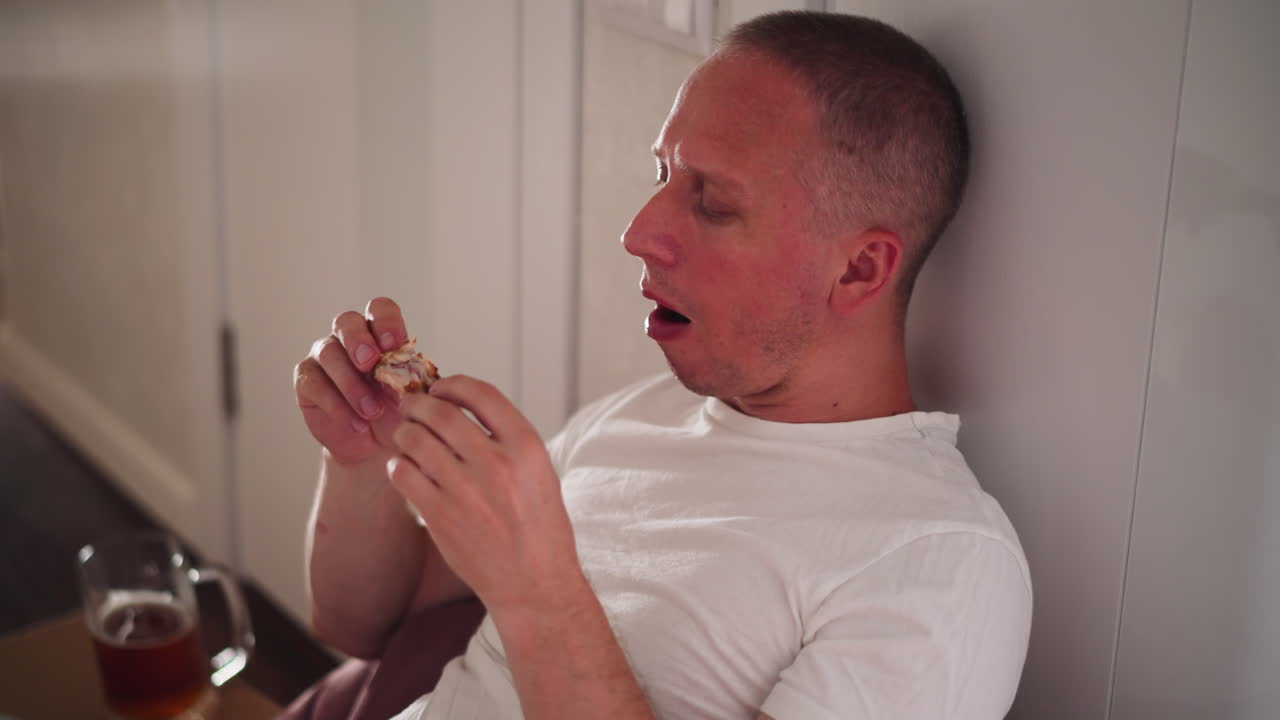 white man wearing casual white t shirt relaxing indoors eating chicken meat passionately while seated on bean bag beside table with glass of beer