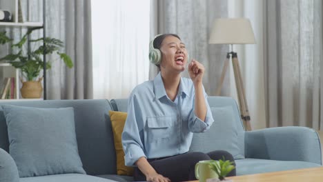 asian woman with headphones listening to music and singing while sitting on sofa in the living room