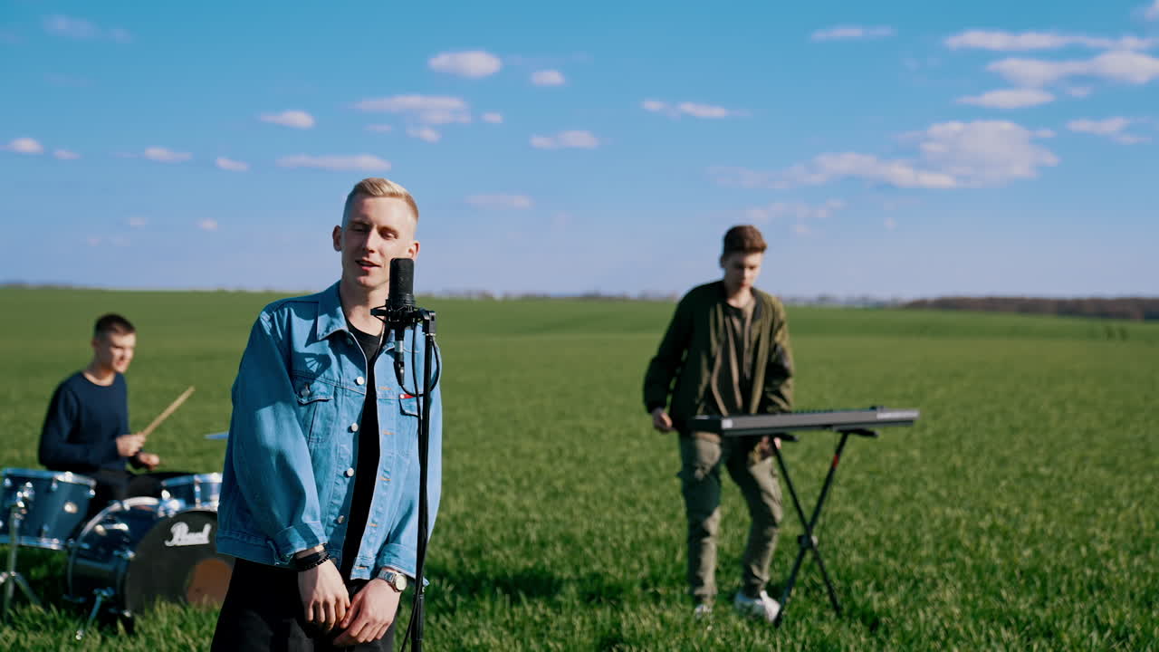 Music group performance in open air. Blond guy soloist singing into microphone while musicians playing the acoustic guitar and drums on field background.