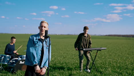 music group performance in open air. blond guy soloist singing into microphone while musicians playing the acoustic guitar and drums on field background.