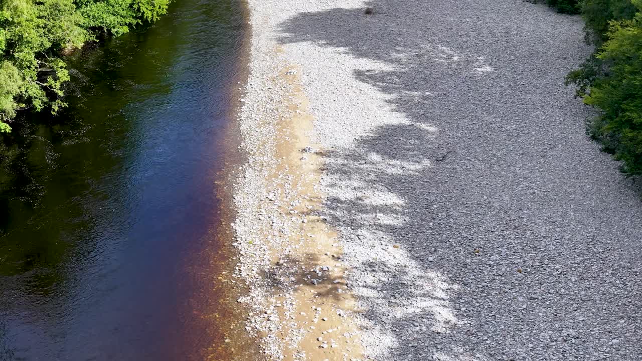 Drone glides above clear river, stony bank, and lush forest in bright natural daylight